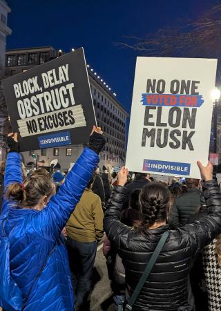 Protesters gather at night holding signs. One reads 'BLOCK, DELAY, OBSTRUCT – NO EXCUSES,' and another says 'NO ONE VOTED FOR ELON MUSK,' both with 'Indivisible' branding. A crowd fills the street in front of illuminated buildings.