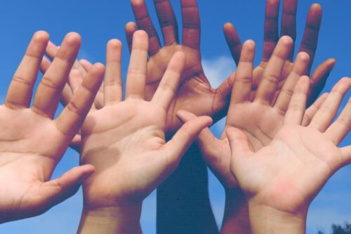 Photo of five hands of various skin tones raised up in the air with fingers spread wide.