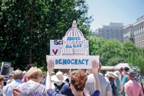 Person holding protest sign with the state capital as the background. The words “Above All Else Democracy” written on top of sign.
